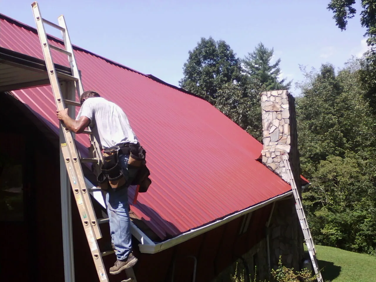 Roofer on a ladder beside a red metal roof and stone chimney — Soffit & Fascia Repair by Roofing Brothers in Fingal